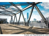 The image is a photograph of a wooden bridge with a view of a city skyline in the background. The bridge is made of steel and...