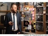 The image shows a man standing in front of a cork board covered in various photographs and post-it notes. He is wearing a dar...