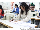 The image shows a young woman working on a sewing machine in a workshop. She is wearing a white sweater and has long curly ha...