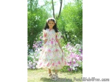 The image shows a young girl standing in a garden with a beautiful floral dress. She is wearing a white dress with pink and y...