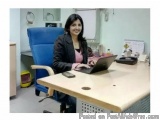 The image shows a young woman sitting at a desk in an office. She is wearing a black blazer and a pink shirt, and is smiling...