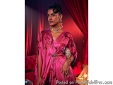 The image is a portrait of a woman wearing a traditional Indian outfit. She is standing in front of a red curtain with a chan...