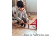 The image shows a young boy playing with a wooden toy train set on the floor. He is sitting on the wooden floor and is focuse...
