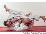 The image shows two men in a martial arts (BJJ) fight. They are both wearing white judo uniforms and are in the middle of a s...