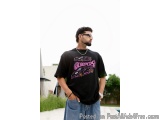 The image shows a young man standing in a garden with his hands in his pockets. He is wearing a black t-shirt with a graphic...