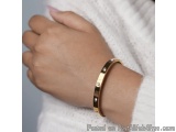 The image shows a close-up of a person's wrist wearing a gold bangle bracelet. The bracelet is made of a thin, gold-colored m...