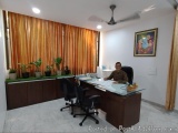 The image shows an office space with a large wooden desk in the center. The desk is made of wood and has a glass top. There a...