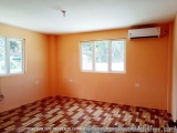 The image shows an empty room with orange walls and a wooden floor. The room has two windows on the left wall, one on the rig...