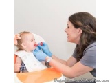 The image shows a young woman sitting in a high chair with a baby in her lap. The baby is wearing a white bib and is being ex...