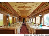 The image shows the interior of a luxurious dining car. The car has a high ceiling with intricate gold designs and a red carp...