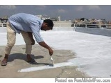 The image shows a man working on a flat roof. He is wearing a blue shirt and khaki pants and is bending down to pick up a whi...