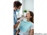 The image shows a young woman sitting in a dental chair with her mouth open, smiling widely. She is wearing a white t-shirt a...