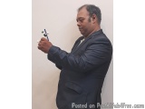 The image shows a middle-aged man in a dark blue suit standing in front of a white wall. He is holding a small model of a mol...