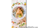 The image is a photograph of a baby girl sitting in a bathtub. The bathtub is covered with a white cloth and is decorated wit...