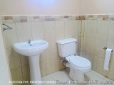 The image shows a small bathroom with a white pedestal sink and a white toilet. The walls are covered in beige tiles with sma...