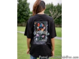 The image shows a woman standing in a park with her back to the camera. She is wearing a black t-shirt with a graphic design...