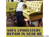 The image shows a man working on a sofa upholstery repair in a workshop. He is standing in front of a large yellow sofa that...