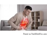 The image shows a young man sitting on a couch in a living room. He is wearing a beige sweater and appears to be in pain or d...