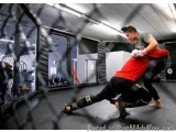 The image shows two men engaged in a martial arts fight inside a cage. The cage is made of metal mesh and has a high ceiling...
