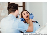 The image shows a young woman sitting in a dental chair with her mouth open, while a dentist is examining her teeth with a de...