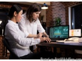 The image shows two women sitting at a desk in an office, working on a computer. They are both wearing white shirts and appea...