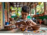 The image shows a young boy sitting at a table in a pottery studio. He is wearing a blue t-shirt and has short dark hair. The...