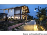 The image shows a modern two-story house with a white exterior and a black roof. The house is located on a hillside with tree...