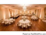 The image shows a large ballroom with a high ceiling and a chandelier hanging from it. The room is decorated with white table...