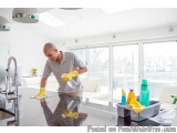 The image shows a man in a kitchen, wearing a grey t-shirt and yellow rubber gloves, cleaning the countertop with a rag and a...