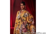 The image shows a woman standing in front of a red curtain. She is wearing a long, flowing yellow dress with a floral print i...