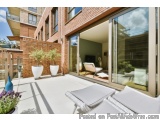 The image shows a modern outdoor patio area of a brick building. The patio is made of concrete and has two white lounge chair...