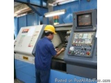 The image shows a man wearing a yellow hard hat and blue overalls operating a large industrial machine in a factory. The mach...
