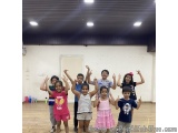 The image shows a group of children standing in a dance studio with their hands up in the air. They are all smiling and appea...
