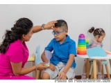 The image shows a young boy sitting on a chair in a classroom. He is wearing a blue shirt and glasses and is looking at a sta...