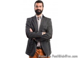 The image is a portrait of a man standing with his arms crossed in front of him. He is wearing a grey blazer over a white shi...