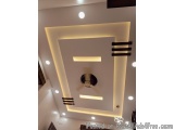 The image shows a modern ceiling design in a room. The ceiling is white and has a square shape with a black circular light fi...