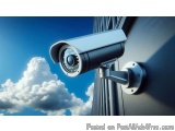 The image shows a security camera mounted on the side of a building. The camera is round and has a silver body with a black l...