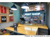 The image shows the interior of a print shop. The shop has a wooden counter with a computer monitor and a keyboard on it. Abo...