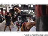 The image shows a young man in a martial arts gym, kicking a punching bag. He is wearing a black and red martial arts uniform...
