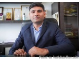The image is a portrait of a man sitting at a desk in an office. He is wearing a dark blue blazer and a light blue shirt. He...