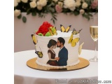 The image is of a wedding cake on a table. The cake is white with a gold base and is decorated with a red rose and yellow but...