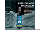 The image shows a bottle of Sushain Pure Magnesium Oil Spray on a bedside table next to an alarm clock. The bottle is black w...