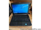 The image shows a black laptop computer sitting on top of a wooden desk. The laptop is open and the screen is turned on, disp...