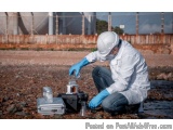 The image shows a person wearing a white hard hat, a white lab coat, and blue gloves, kneeling on the ground and working on a...