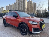The image shows a red Audi Q7 SUV parked on a gravel road in an urban area. The car is facing towards the left side of the im...