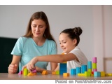 The image shows a mother and daughter playing with colorful building blocks on a table. The mother is wearing a light blue t-...