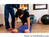 The image shows a man in a black t-shirt and blue pants performing a push-up exercise on a blue yoga mat. He is holding two o...