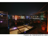 The image shows an outdoor patio area at night. The patio is covered with a wooden pergola and has several potted plants on t...