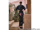 The image shows a woman standing on a set of stairs in a garden. She is wearing a black jumpsuit with a floral print in shade...