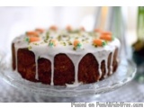 The image is of a round cake on a glass cake stand. The cake appears to be a carrot cake with a golden brown color and a driz...
